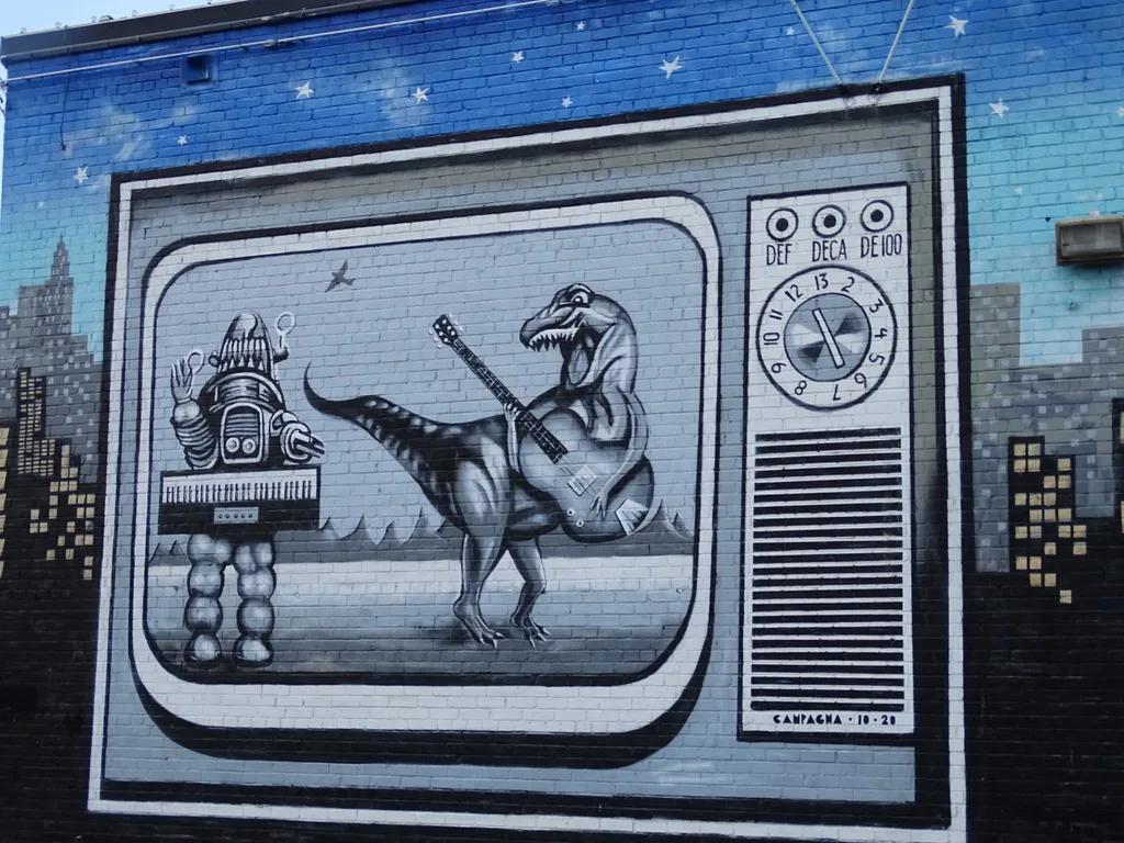 A mural in Dallas of a vintage television set, painted on brick: a rounded screen in a panel, three knobs and a numbered dial down the right side, a grille below them, and a robot and a guitar-playing dinosaur on the screen.